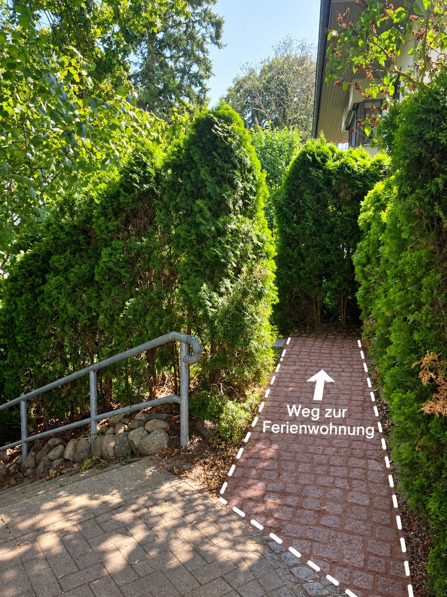 Gepflasterter Weg zwischen Hecken mit Pfeil und der Aufschrift „Weg zur Ferienwohnung“